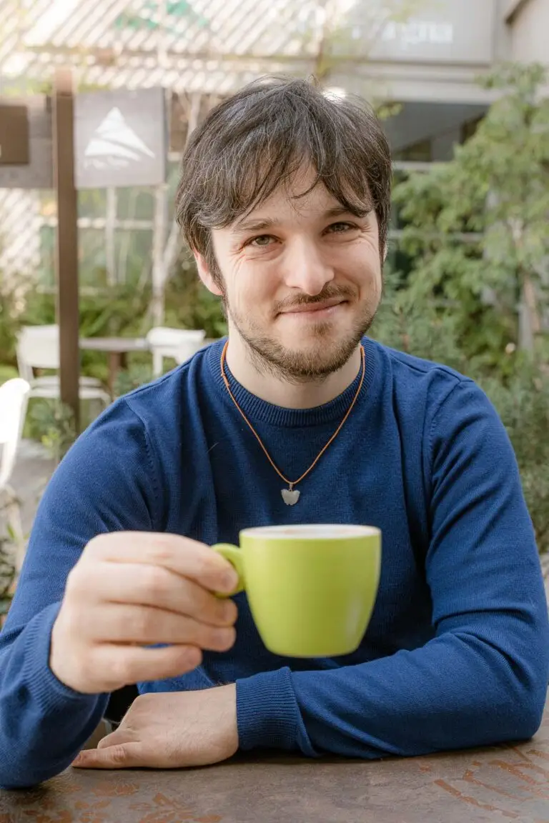 Davide riccio, porge una tazza di caffè a chi guarda la foto. Indossa una felpa blu scuro, un ciondolo bianco. La tazza è verde chiaro.